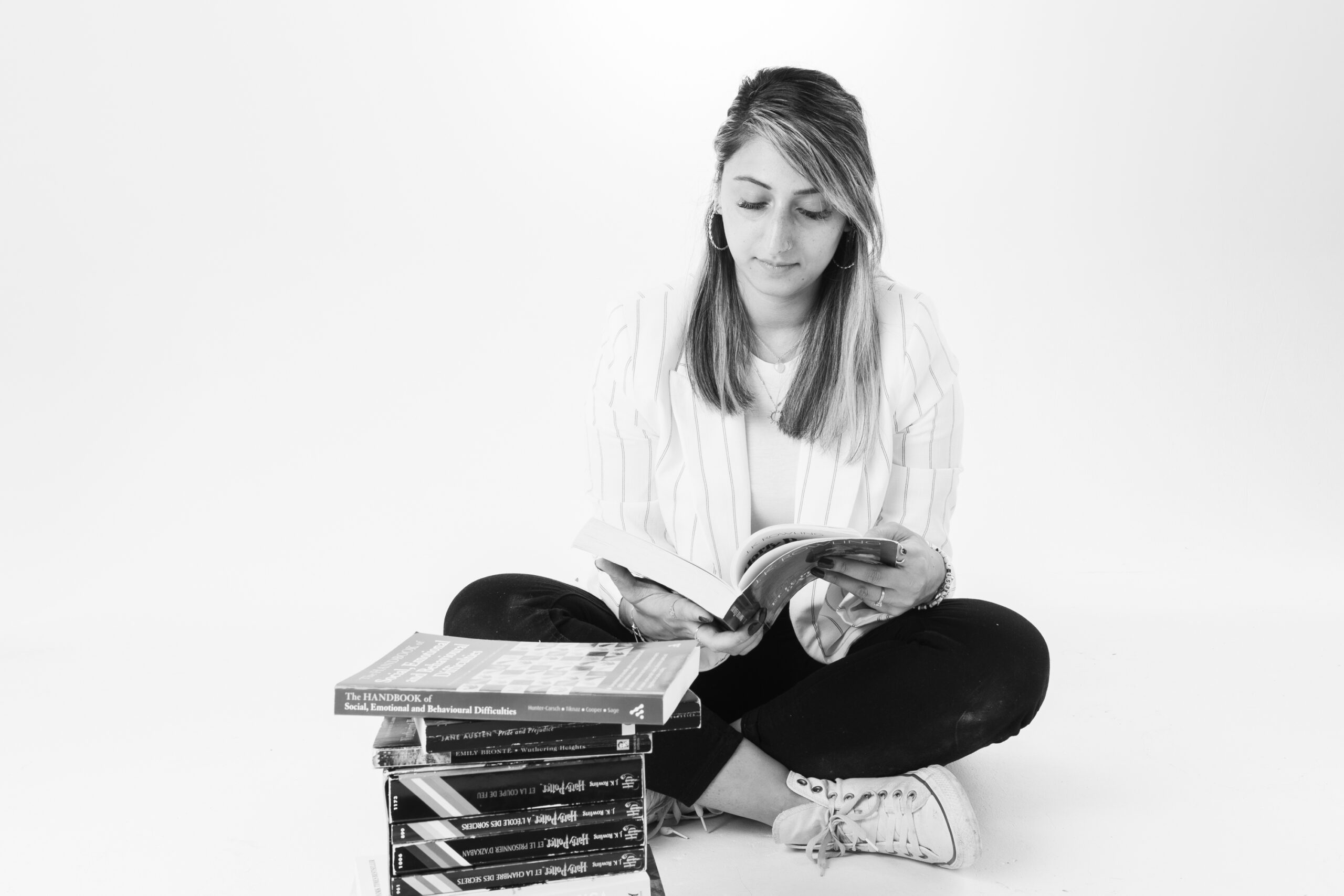 The founder of the business who is also a private SEN tutor, looks down at a book, cross legged. The image is in black and white. She is from South Asian origin and has a stack of books in front of her.